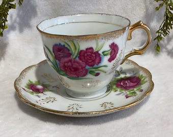 Puede incluir: Un juego de taza y platillo de té vintage. La taza y el platillo de porcelana blanca están adornados con un diseño floral de claveles rosa intenso y ribetes dorados. La taza tiene un asa dorada y el platillo tiene un borde festoneado.