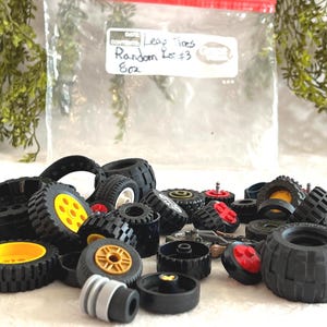 May include: A collection of black Lego tires with yellow, red, and white accents. The tires vary in size and tread pattern. A plastic bag in the background has the text "Leaf Tires Random Lot #3 8oz."