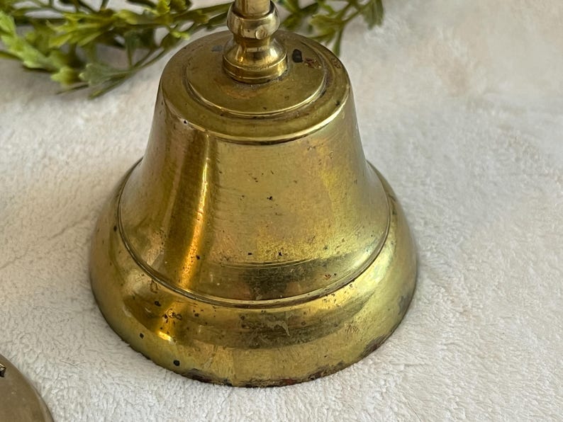 May include: A vintage brass bell with a rounded base and a handle. The bell has a tarnished, aged appearance, with visible wear and tear. The bell is likely used for decorative purposes or as a collectible item.