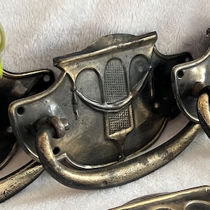 May include: Antique bronze-toned drawer pulls with a decorative arched design and curved handles. The pulls have a detailed, textured surface and are likely made of metal. These vintage-style pulls are suitable for furniture restoration projects.