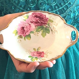 May include: A small, off-white dish with a scalloped edge and two handles. The dish is decorated with pink roses and green leaves. The rim is trimmed with gold. A vintage-style serving dish.