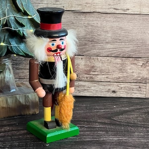 Op de afbeelding: Een houten notenkrakerfiguur met een zwarte hoge hoed met een rode band, een zwart jasje en een gele broek. Het figuur heeft wit haar en een baard, een rode neus en een zwarte snor. Het houdt een geel koord vast met een bruin pluizig accessoire.