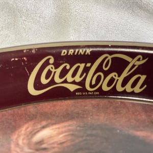 Może przedstawiać: Zabytkowy taca do serwowania Coca-Cola z bordowym obramowaniem i złotym logo Coca-Cola. Taca ma napis "DRINK" nad logo i "REG. U.S. PAT. OFF." pod nim. Taca ma klasyczny design.