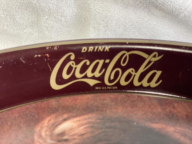 Może przedstawiać: Zabytkowy taca do serwowania Coca-Cola z bordowym obramowaniem i złotym logo Coca-Cola. Taca ma napis "DRINK" nad logo i "REG. U.S. PAT. OFF." pod nim. Taca ma klasyczny design.