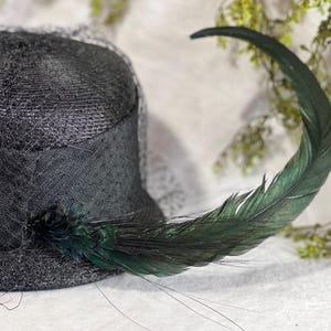 May include: A black hat with a veil and a large, curved green feather. The hat is made of a woven material and has a dark, elegant appearance. The feather adds a touch of flair.