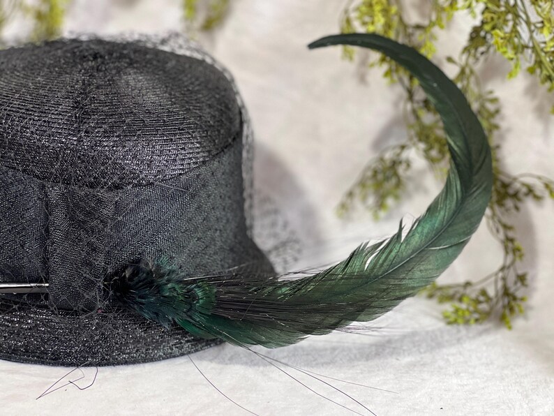 May include: A black hat with a veil and a large, curved green feather. The hat is made of a woven material and has a dark, elegant appearance. The feather adds a touch of flair.