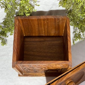 May include: A square wooden box with a lid, featuring a warm brown color and visible wood grain. The box has carved details on the front and is open, revealing the interior. Decorative greenery is in the background.