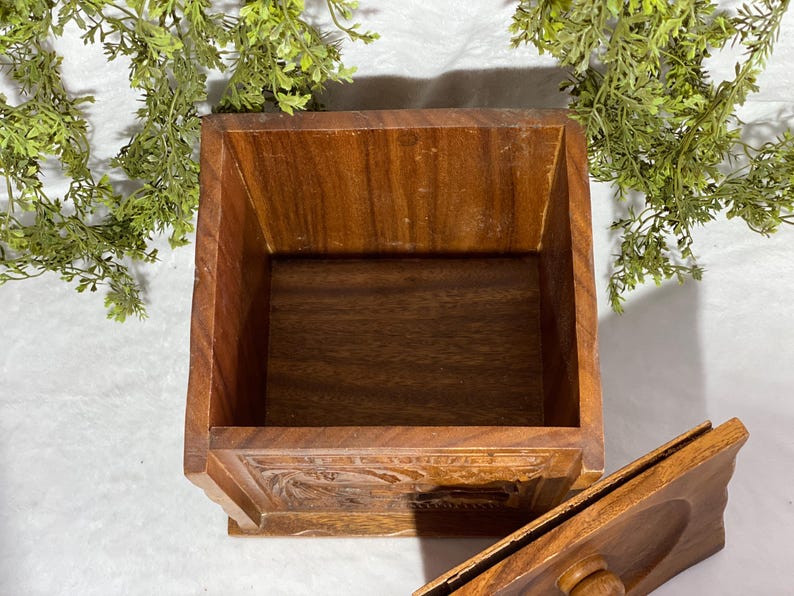 May include: A square wooden box with a lid, featuring a warm brown color and visible wood grain. The box has carved details on the front and is open, revealing the interior. Decorative greenery is in the background.