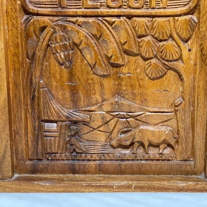 May include: A wooden box with intricate carvings depicting a tropical landscape. The relief carving shows palm trees, a hut, a water buffalo, and a person. The box is made of a warm, medium-toned wood.