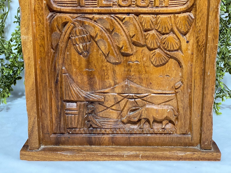 May include: A wooden box with intricate carvings depicting a tropical landscape. The relief carving shows palm trees, a hut, a water buffalo, and a person. The box is made of a warm, medium-toned wood.