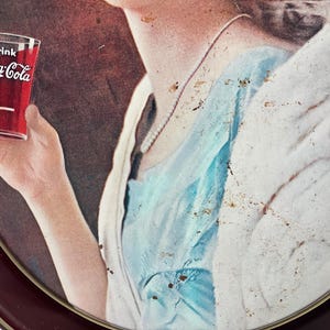 Może przedstawiać: Reklama Coca-Cola w stylu vintage przedstawiająca szklankę napoju. Szklanka jest trzymana przez osobę, z widocznym logo Coca-Cola. Obraz ma estetykę retro, z paletą kolor&oacute;w czerwonego, białego i niebieskiego.