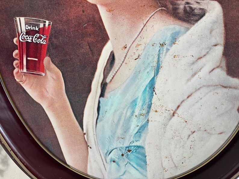 Może przedstawiać: Reklama Coca-Cola w stylu vintage przedstawiająca szklankę napoju. Szklanka jest trzymana przez osobę, z widocznym logo Coca-Cola. Obraz ma estetykę retro, z paletą kolor&oacute;w czerwonego, białego i niebieskiego.