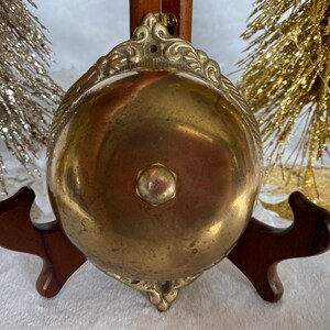 Op de afbeelding: Een messing bel met een sierlijk ontwerp en een houten voet. De bel is gepolijst en heeft een warme, gouden kleur.