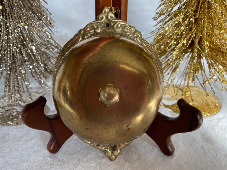 Op de afbeelding: Een messing bel met een sierlijk ontwerp en een houten voet. De bel is gepolijst en heeft een warme, gouden kleur.