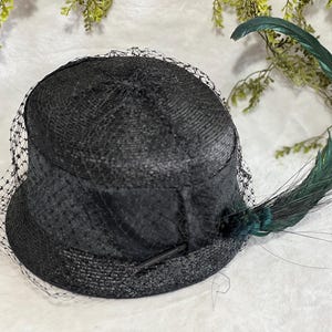 May include: A black hat with a rounded crown and a short brim, adorned with a delicate black netting veil. A long, curved green feather adds a striking accent. The hat is likely vintage or inspired by vintage styles.
