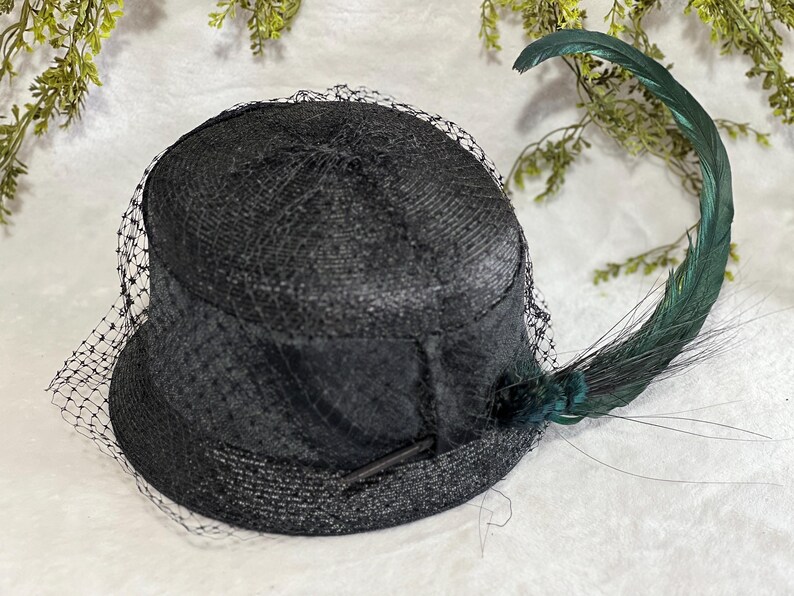 May include: A black hat with a rounded crown and a short brim, adorned with a delicate black netting veil. A long, curved green feather adds a striking accent. The hat is likely vintage or inspired by vintage styles.