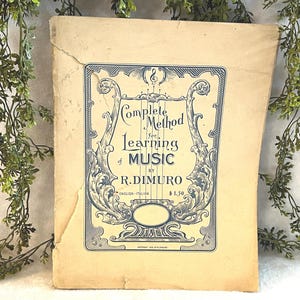 May include: A vintage music book titled "Complete Method for Learning of Music" by R. DiMuro. The cover is a light beige with a decorative blue border and text. The book is slightly worn, suggesting its age.