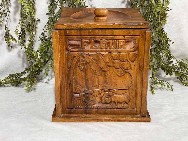 May include: A wooden flour container with a lid and a carved design. The word "FLOUR" is carved across the top. The container has a rectangular shape and a detailed relief carving of a rural scene.