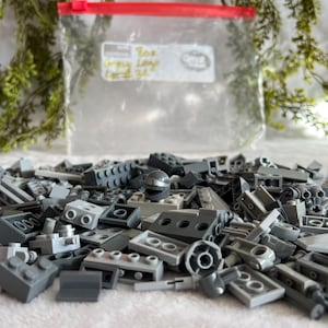 Puede incluir: Un conjunto de ladrillos Lego grises de varias formas y tamaños, incluyendo baldosas planas, bloques y piezas especializadas. Una bolsa de plástico transparente con cierre rojo está en el fondo, con texto escrito a mano. Los ladrillos están esparcidos sobre una superficie blanca.