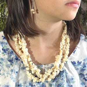 May include: A cream-colored, layered necklace with matching earrings. The necklace is made of small, flower-shaped beads. The person is wearing a sleeveless top with a blue and white floral pattern. The background includes green foliage.