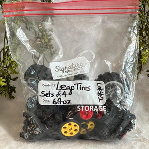 Op de afbeelding: Een doorzichtige plastic zak gevuld met zwarte Lego-banden, sommige met rode en gele accenten. De zak heeft een rode rits en een wit label met de tekst "Lego Tires, Sets of 4, 181g". De zak is gelabeld met "STORAGE".
