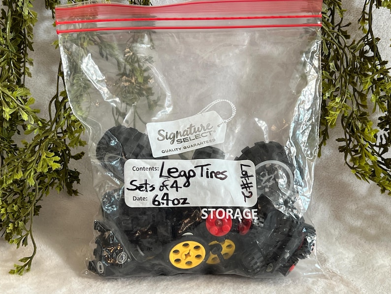 Op de afbeelding: Een doorzichtige plastic zak gevuld met zwarte Lego-banden, sommige met rode en gele accenten. De zak heeft een rode rits en een wit label met de tekst "Lego Tires, Sets of 4, 181g". De zak is gelabeld met "STORAGE".