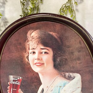 Może przedstawiać: Zabytkowy podkładka do serwowania Coca-Coli z portretem kobiety trzymającej szklankę Coca-Coli. Taca ma ciemnobrązową owalną ramę. Kobieta ma brązowe włosy i ma na sobie jasnoniebieską i białą sukienkę.