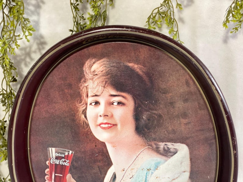 Może przedstawiać: Zabytkowy podkładka do serwowania Coca-Coli z portretem kobiety trzymającej szklankę Coca-Coli. Taca ma ciemnobrązową owalną ramę. Kobieta ma brązowe włosy i ma na sobie jasnoniebieską i białą sukienkę.