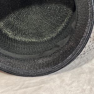May include: A black hat with a wide brim and a black ribbon band. The hat has a textured weave and a delicate black netting veil. The hat is a classic accessory.