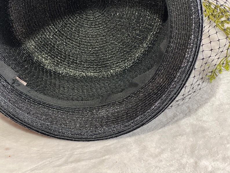 May include: A black hat with a wide brim and a black ribbon band. The hat has a textured weave and a delicate black netting veil. The hat is a classic accessory.