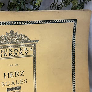 May include: A vintage book cover from Schirmer's Library, featuring the title "Herz Scales" in bold lettering. The cover is a light beige color with a decorative dark green border and ornate detailing. The book is volume 170.