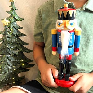 May include: A wooden nutcracker figurine featuring a blue jacket with gold accents, a black crown, and a white beard. The figure stands on a round red base and is painted with blue, red, black, gold, and white colors, showcasing a classic soldier design.