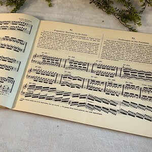 May include: Open book with sheet music and text. The pages are aged with a cream color. The music notes are black and detailed. The text is in English and German. The book is open to a page with musical exercises.