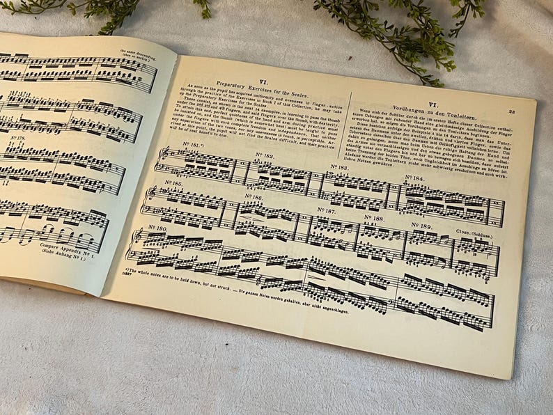 May include: Open book with sheet music and text. The pages are aged with a cream color. The music notes are black and detailed. The text is in English and German. The book is open to a page with musical exercises.