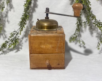 Op de afbeelding: Een vintage houten koffiemolen met een messing bovenkant en een lade. De molen heeft een metalen hendel met een houten knop. Het hout heeft een natuurlijke afwerking en de molen lijkt goed gebruikt. De achtergrond bevat groen.
