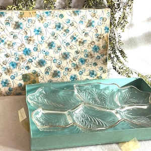 Puede incluir: Una fuente de servir vintage con forma de hoja y borde dorado, en una caja color turquesa. La fuente es de vidrio transparente con detalles de hojas grabadas. La caja tiene una tapa con estampado floral en azul, marrón y crema.