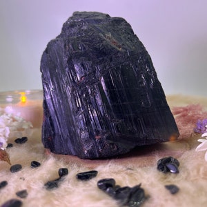 Puede incluir: Un gran cristal de turmalina negra en bruto y sin pulir con estrías. El cristal está sentado sobre una superficie peluda de color marrón claro con pequeños cristales negros esparcidos a su alrededor.