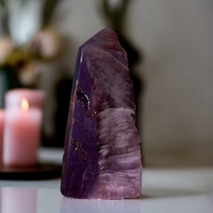 Puede incluir: Una gran punta de cristal púrpura y rosa con una superficie lisa y pulida. El cristal está sobre una superficie blanca.