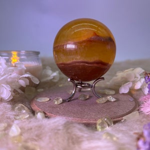 May include: A yellow and brown banded agate sphere on a metal stand. The sphere is polished and has a translucent appearance. The sphere is surrounded by small white and yellow stones.