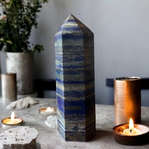 Puede incluir: Una punta de cristal alta, de rayas azules y grises, también conocida como torre u obelisco, se encuentra sobre una superficie gris con dos velas encendidas delante de ella.