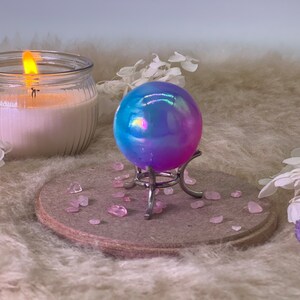 Puede incluir: Una esfera de vidrio azul y morado iridiscente sobre un soporte de metal. La esfera está sentada sobre un posavasos de madera con cristales rosas esparcidos a su alrededor.