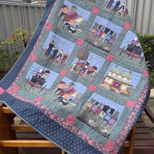 May include: A patchwork quilt featuring a series of printed fabric squares depicting rural farm life, including houses, barns, people in hats, laundry on lines, and cows. The quilt is bordered by dark blue fabric with small white floral patterns.