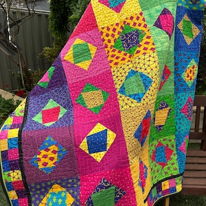 May include: A colourful quilt with a patchwork design featuring various geometric shapes in shades of pink, purple, yellow, green, and blue.
