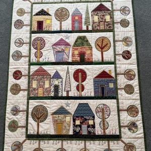May include: A rectangular quilt featuring a grid of houses and trees in various fabric patterns and colours. The design includes four horizontal rows of houses separated by dark green borders, with circular tree motifs along the outer edges.