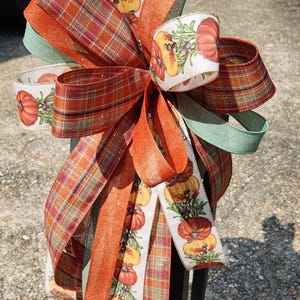 May include: A decorative bow made from multiple ribbons featuring orange and red plaid patterns, solid orange glittery fabric, solid sage green fabric, and a white ribbon printed with a repeating pattern of orange pumpkins and green foliage.