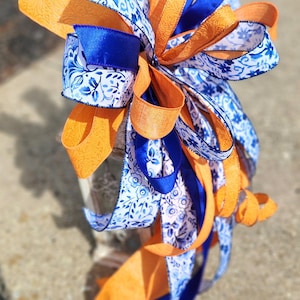 May include: A decorative bow constructed from multiple ribbons featuring a blue and white floral pattern, solid orange textured ribbon, and solid royal blue satin ribbon. The ribbons are arranged in numerous loops and long tails.