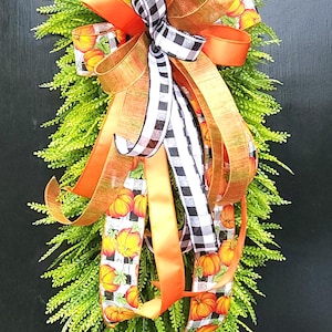 May include: A fall-themed decorative swag featuring a lush green base and a vibrant bow. The bow incorporates orange, gold, and black and white checkered ribbons, with pumpkin patterns. Perfect for autumn decor.
