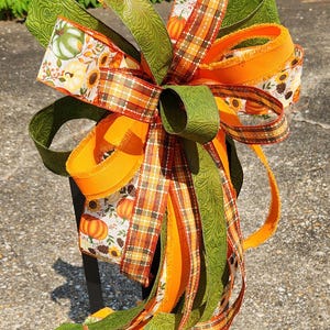 May include: A decorative multi-layered bow featuring a mix of ribbons in solid orange, green with a subtle embossed leaf pattern, and a white print with pumpkins, sunflowers, and pinecones, accented by a brown, orange, and green plaid ribbon.