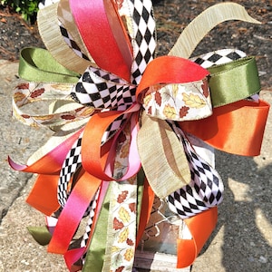 May include: A decorative bow made of multiple ribbons in orange, olive green, hot pink, gold, and a black and white diamond pattern. Some ribbons feature a printed autumn leaf design. The bow is constructed with numerous loops and long trailing ends.