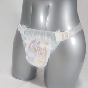 Op de afbeelding: Een wegwerpluier met een cartoonontwerp van twee honden, vastgezet met een doorzichtige plastic riem en witte plastic clips. De luier is ontworpen voor baby's en peuters.